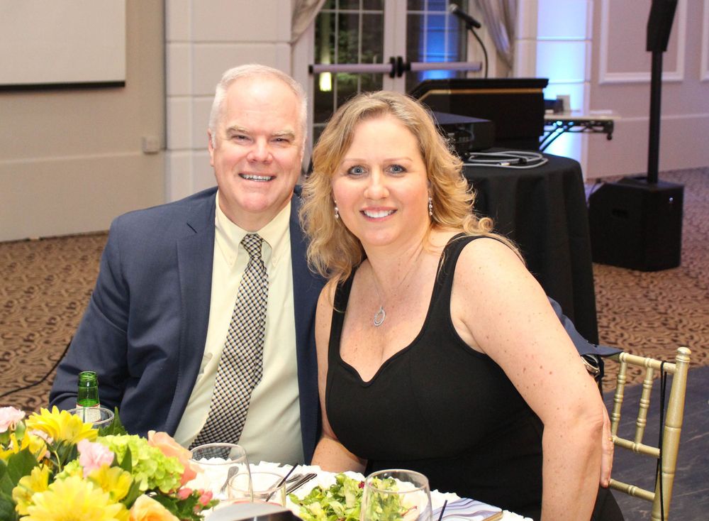 A couple in formal attire seated at an event with yellow floral centerpieces.