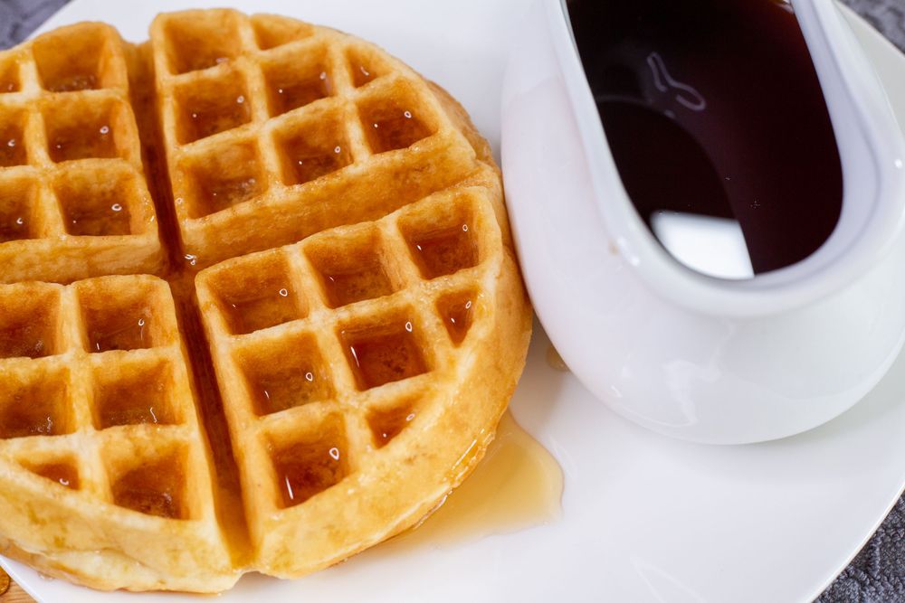 Golden waffle drizzled with syrup next to a cup of dark sauce on a white plate.