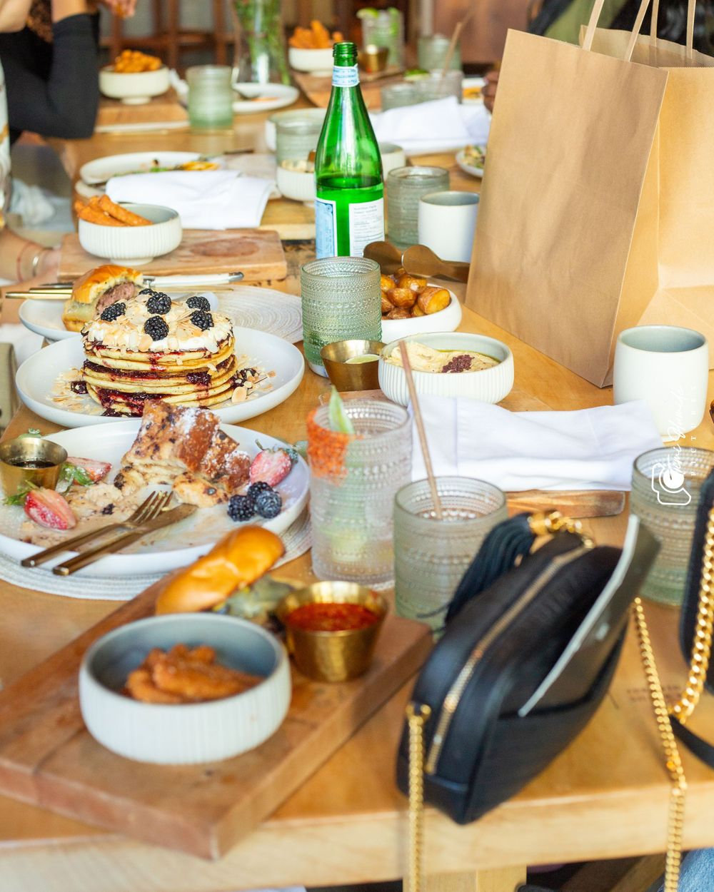Elegant brunch spread featuring blueberry pancakes, drinks, and a black handbag on wooden table.