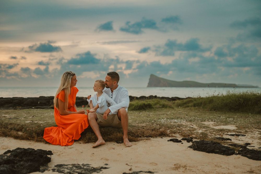 A couple in casual attire sits on a sandy beach at sunset with mountains and ocean in the background.