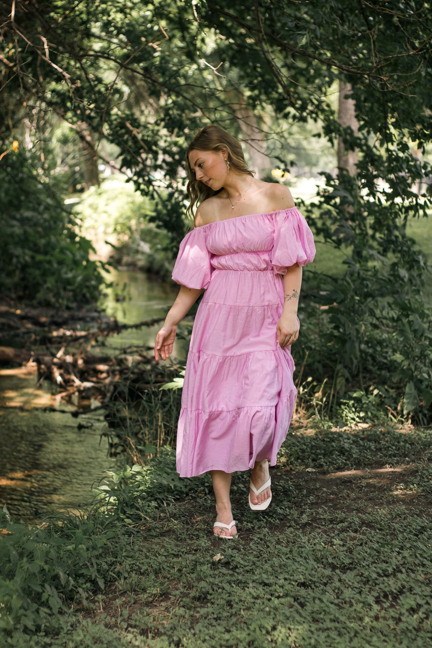 Woman in pink off-shoulder midi dress with puff sleeves walks through lush green forest setting.