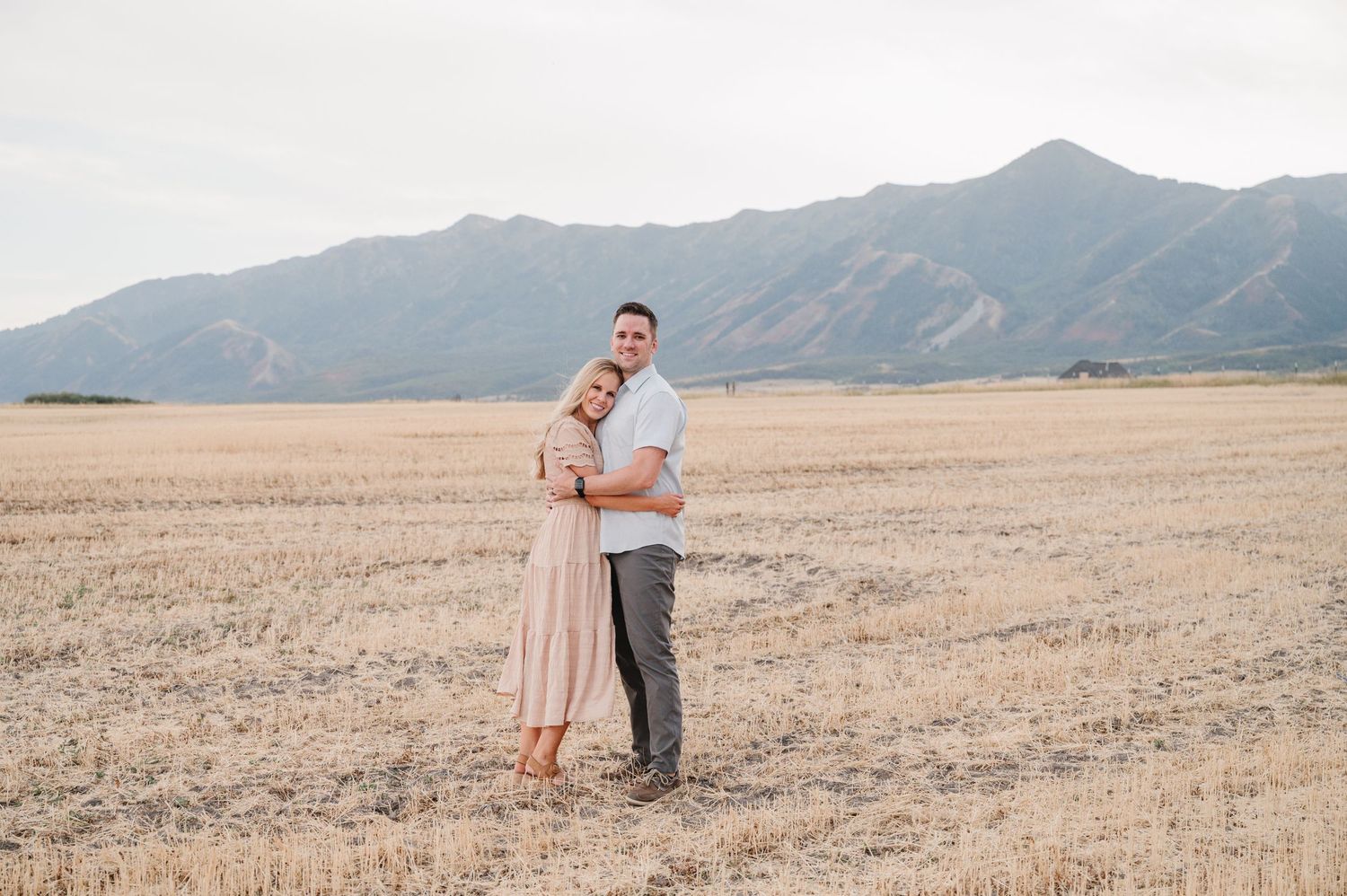 A couple poses together in an open field with mountains in the background at sunset.