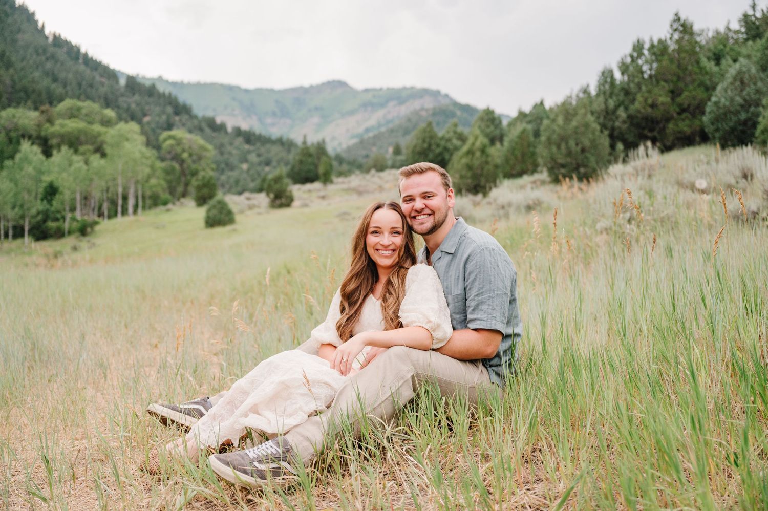 A couple sitting together in tall grass with mountain views in the background.