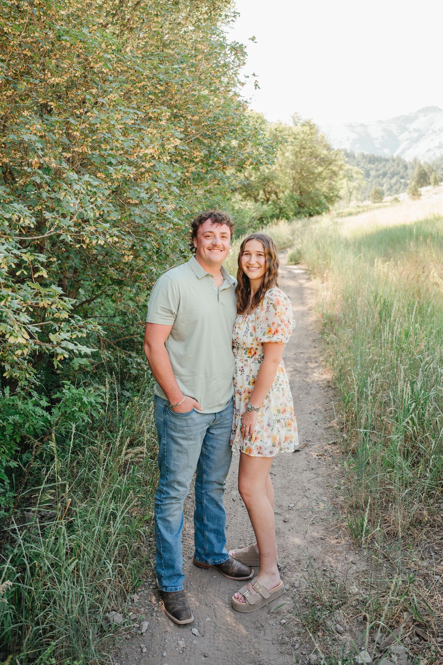 A smiling couple poses on a scenic dirt path surrounded by lush greenery and mountains in soft golden light.