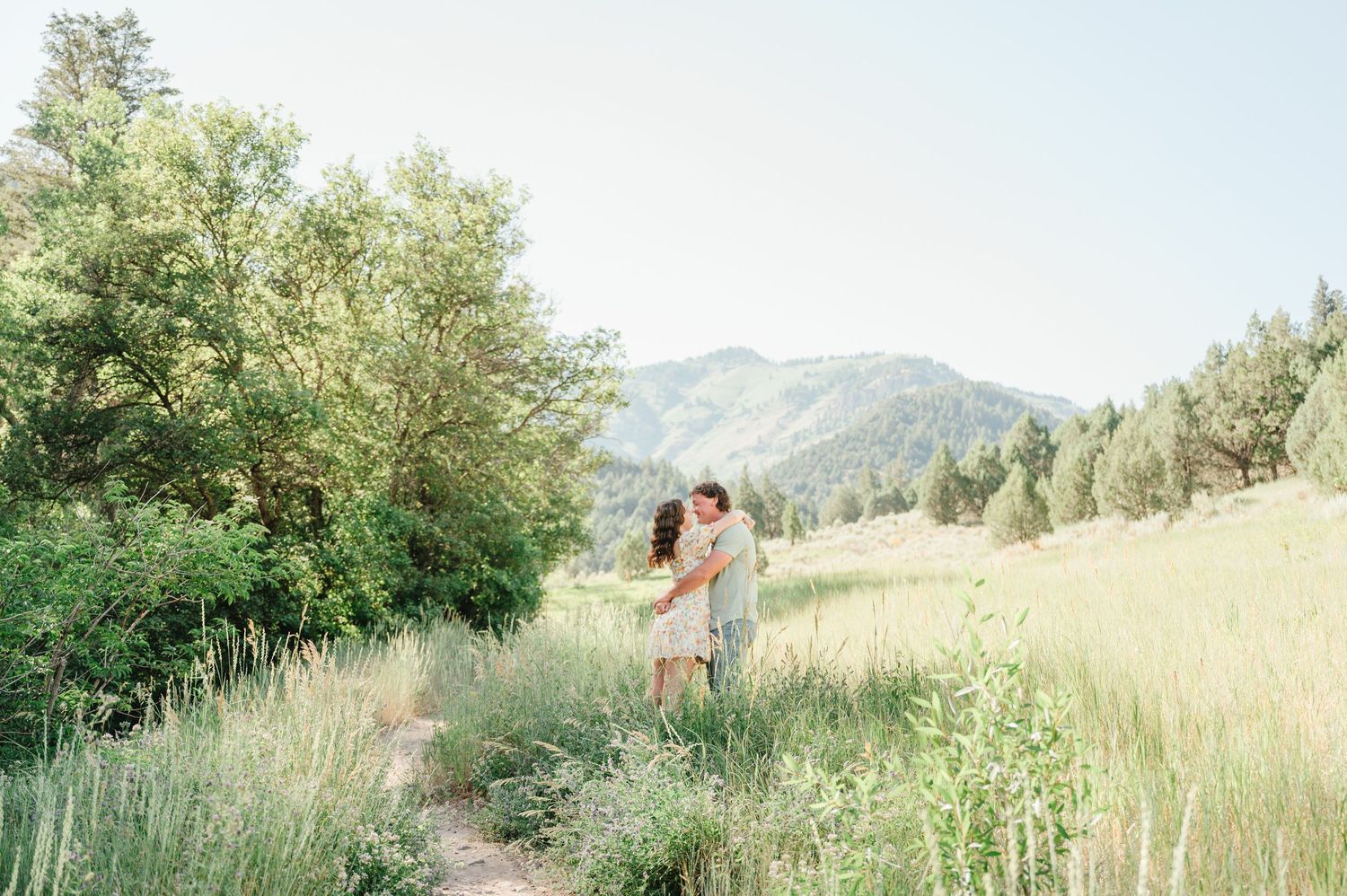 Couple embracing in a lush green meadow with tall grass and mountain views in the background.