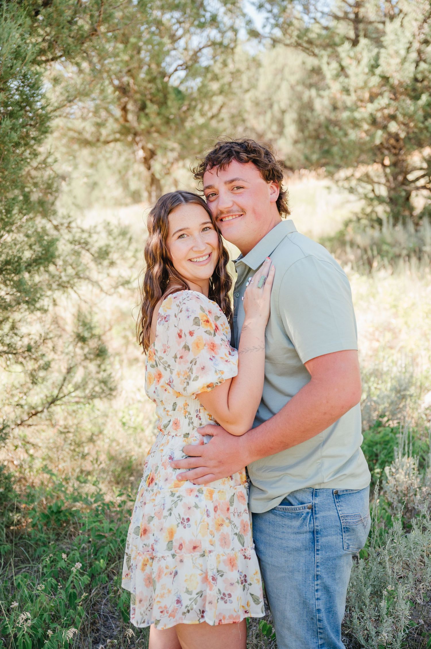 A young couple embraces outdoors in a sunny field, woman in floral dress, man in sage polo and jeans.