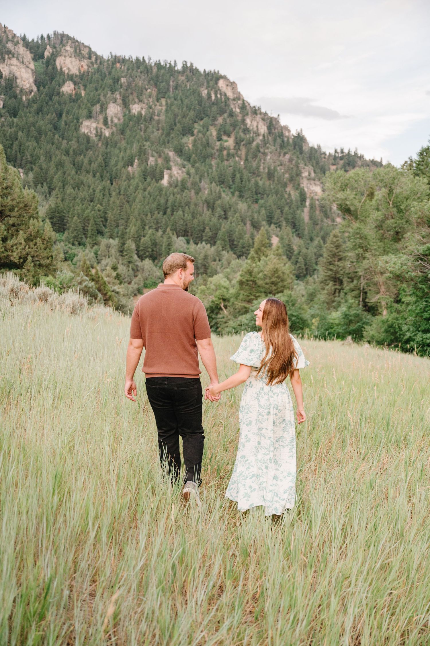 A couple walking hand in hand through tall grass with dramatic mountain cliffs in the background.