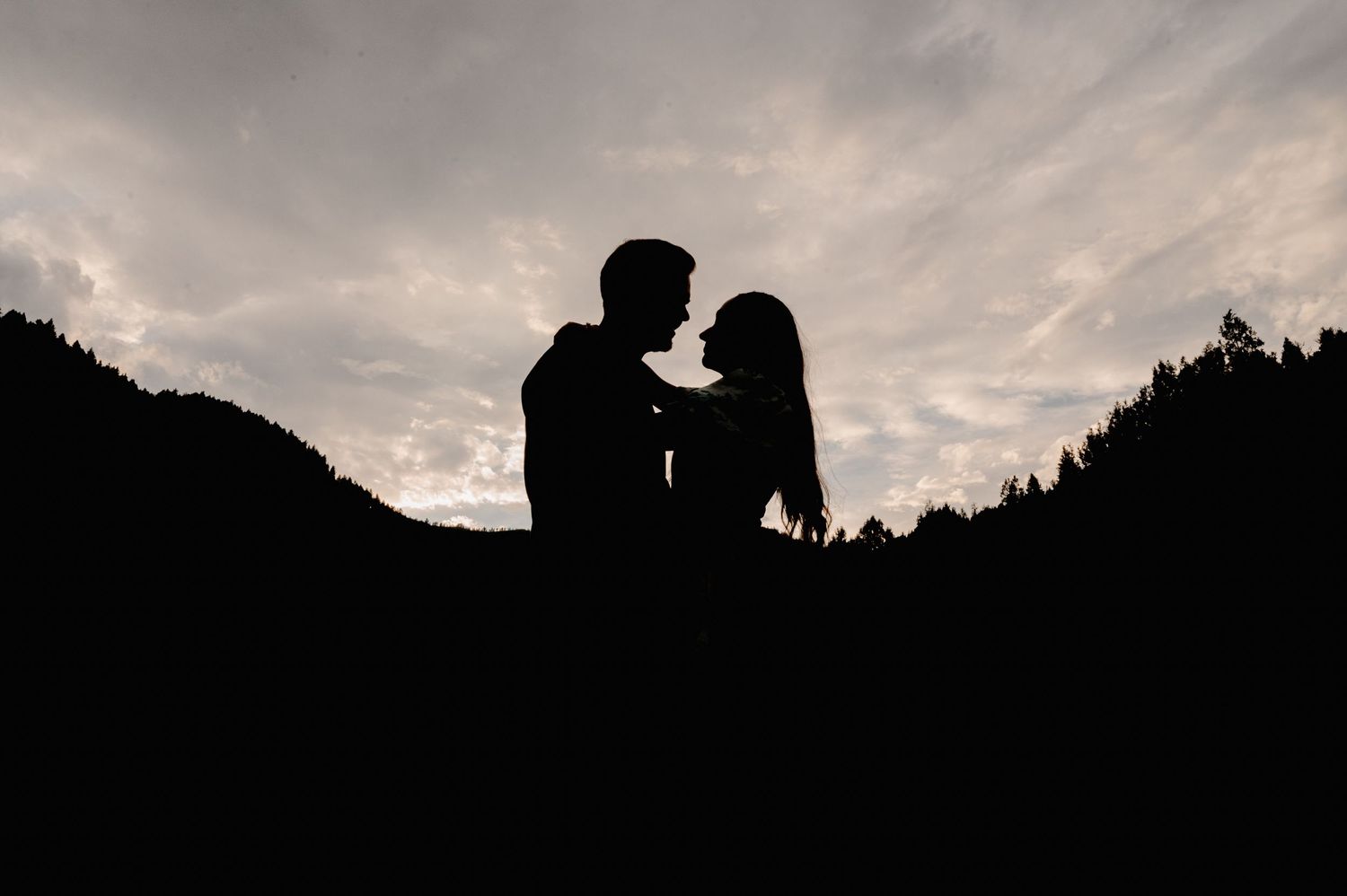 A silhouetted couple against a dramatic sunset sky with mountain ridges in the background.