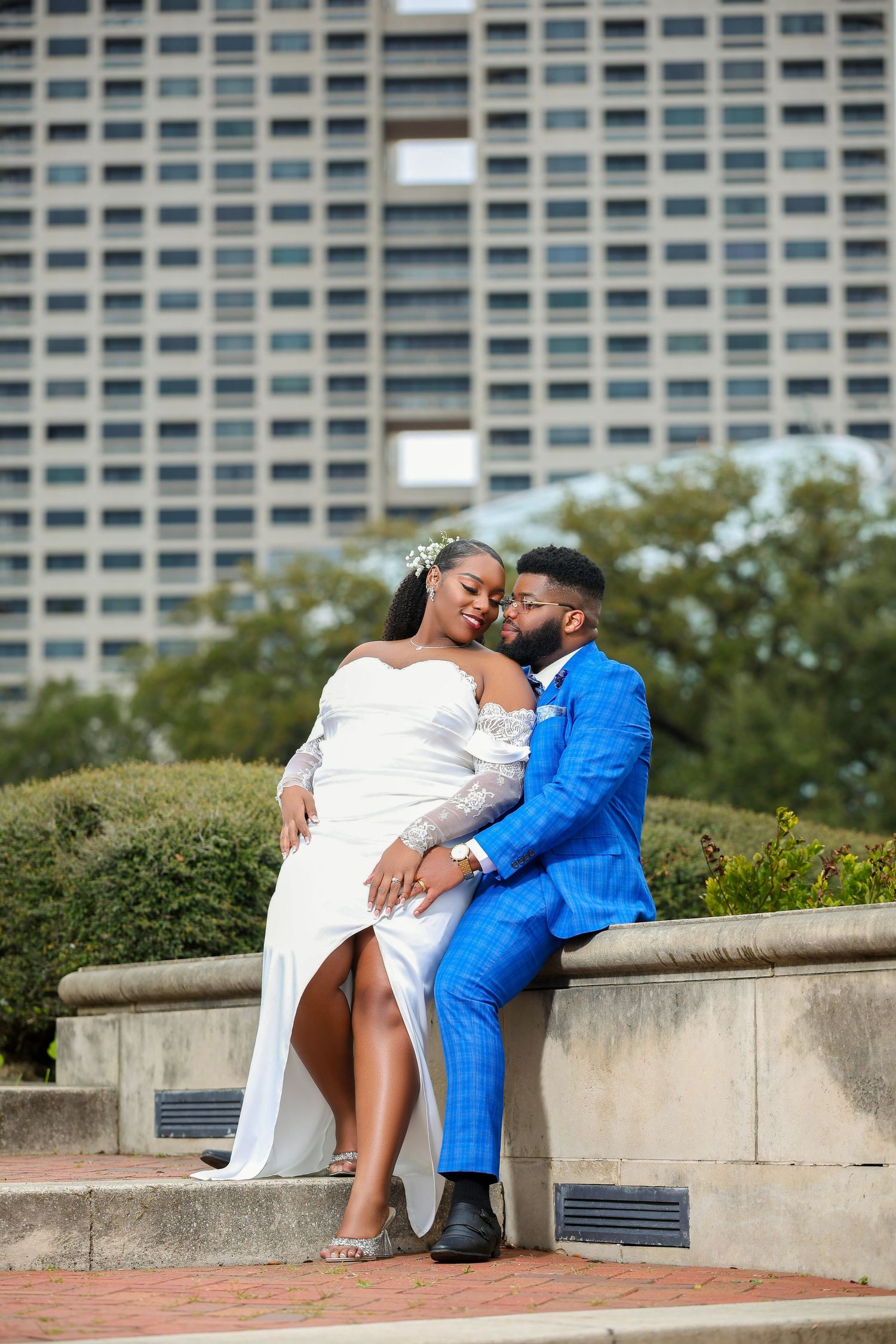 Couple in elegant white dress and bright blue suit embrace while sitting at outdoor urban location near tall building.