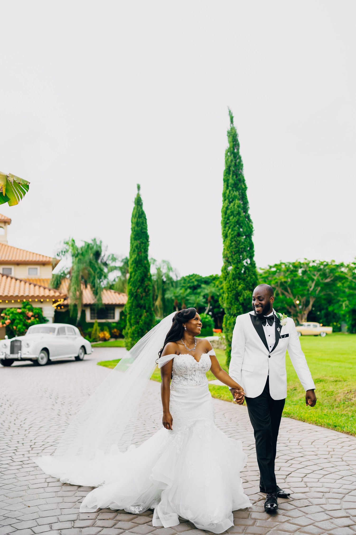 A bride and groom walking hand in hand on a cobblestone path with cypress trees in the background.