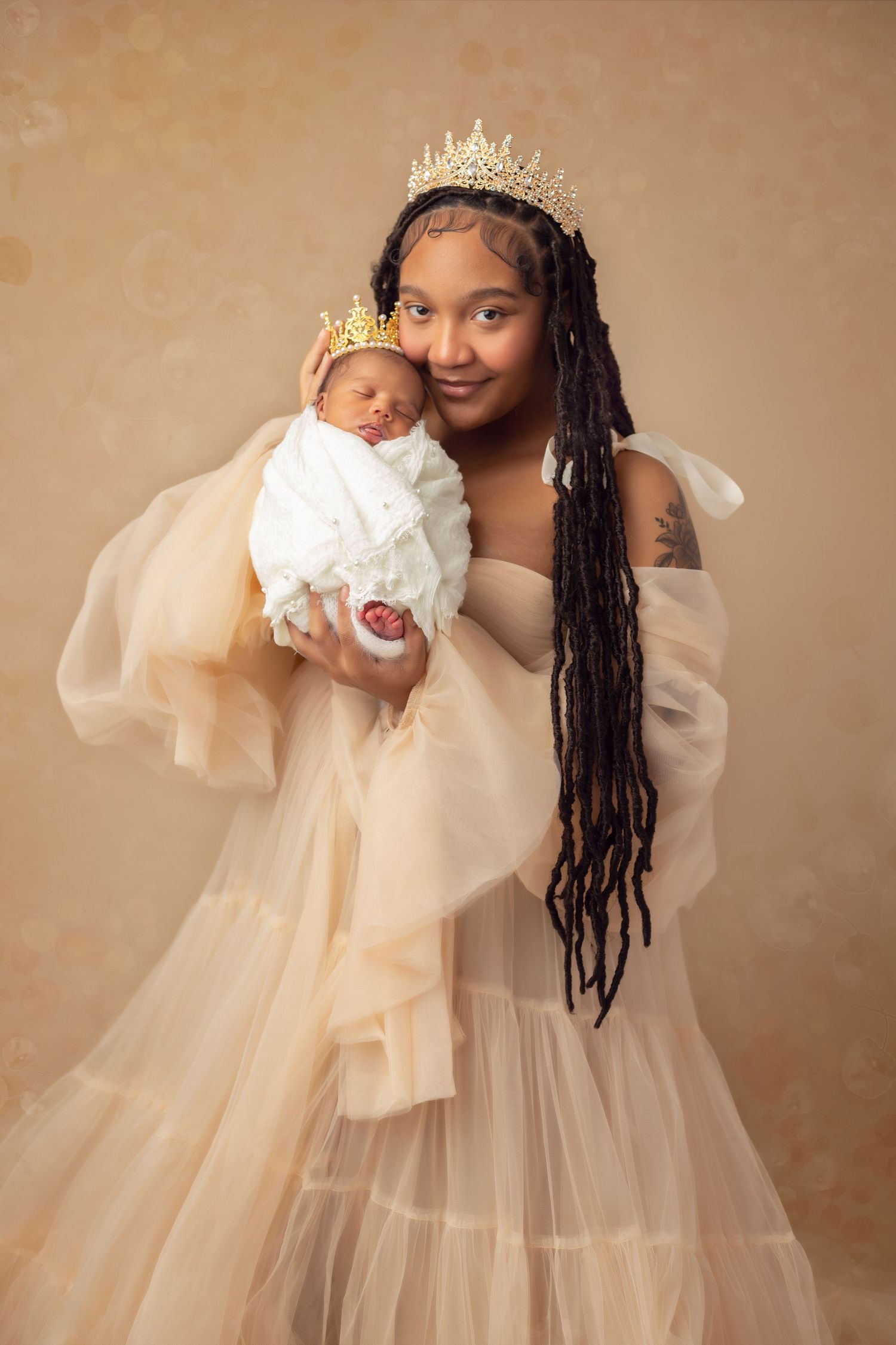Portrait of Mommy and sweet new baby girl both wearing crowns, and a flowing cream color gown against similar color background.