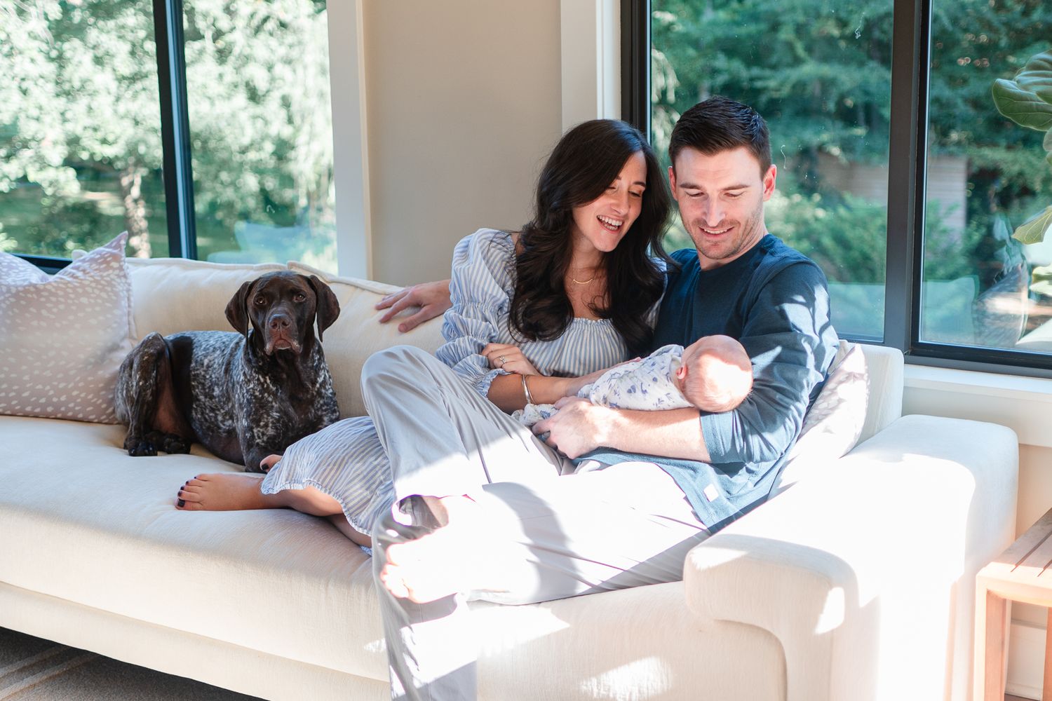 Mom, dad, baby, and family dog enjoy the warmth of sunshine at a newborn session at home.