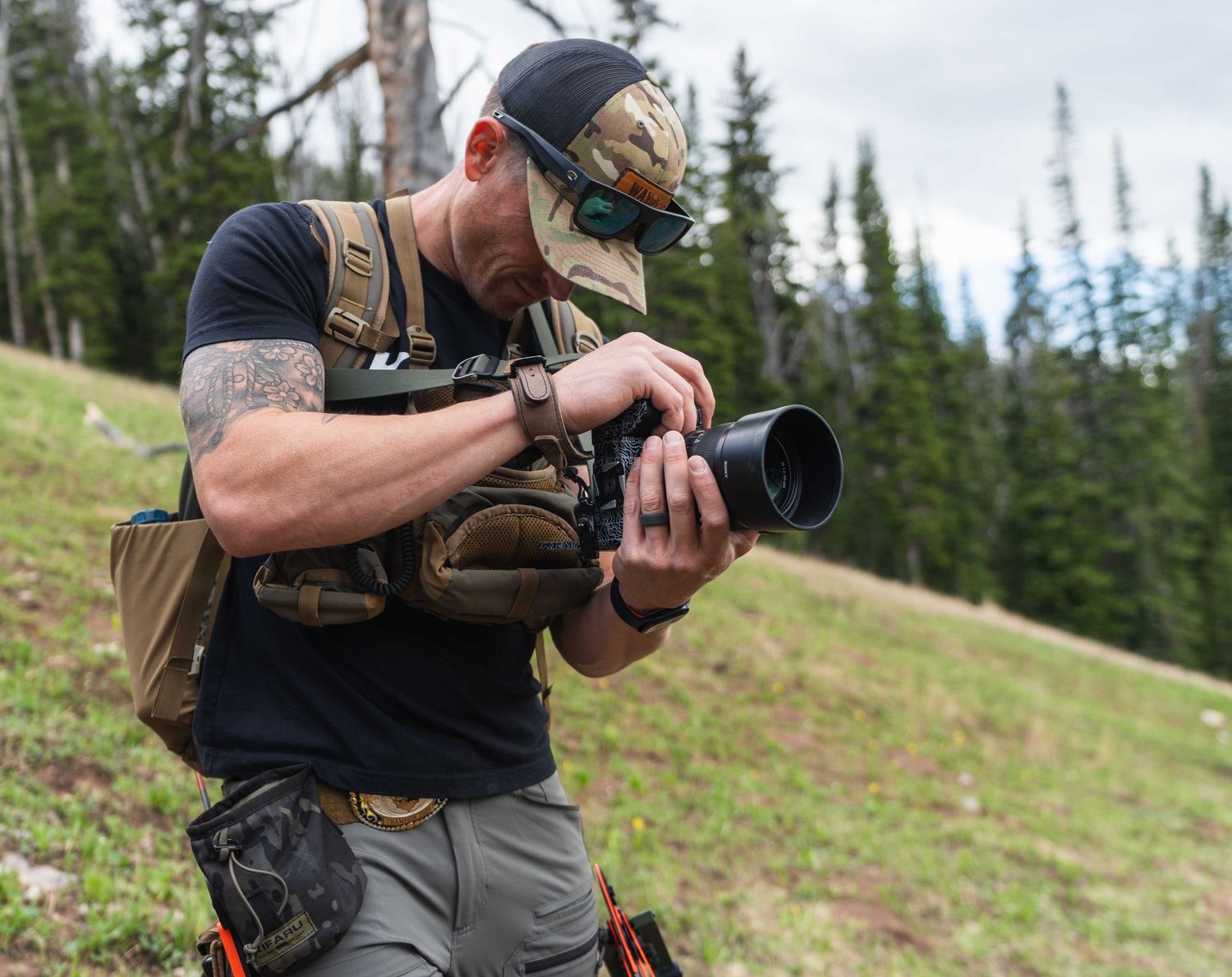 Photographer with camera gear kneeling in mountain meadow with evergreen trees.