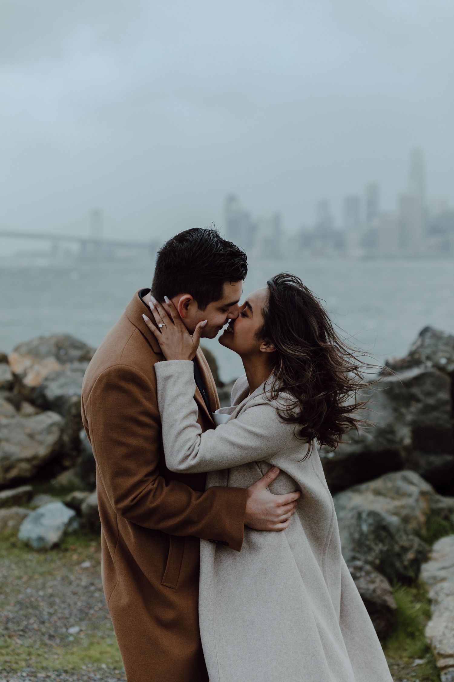 A romantic embrace on a foggy coastline with a city skyline visible in the background.