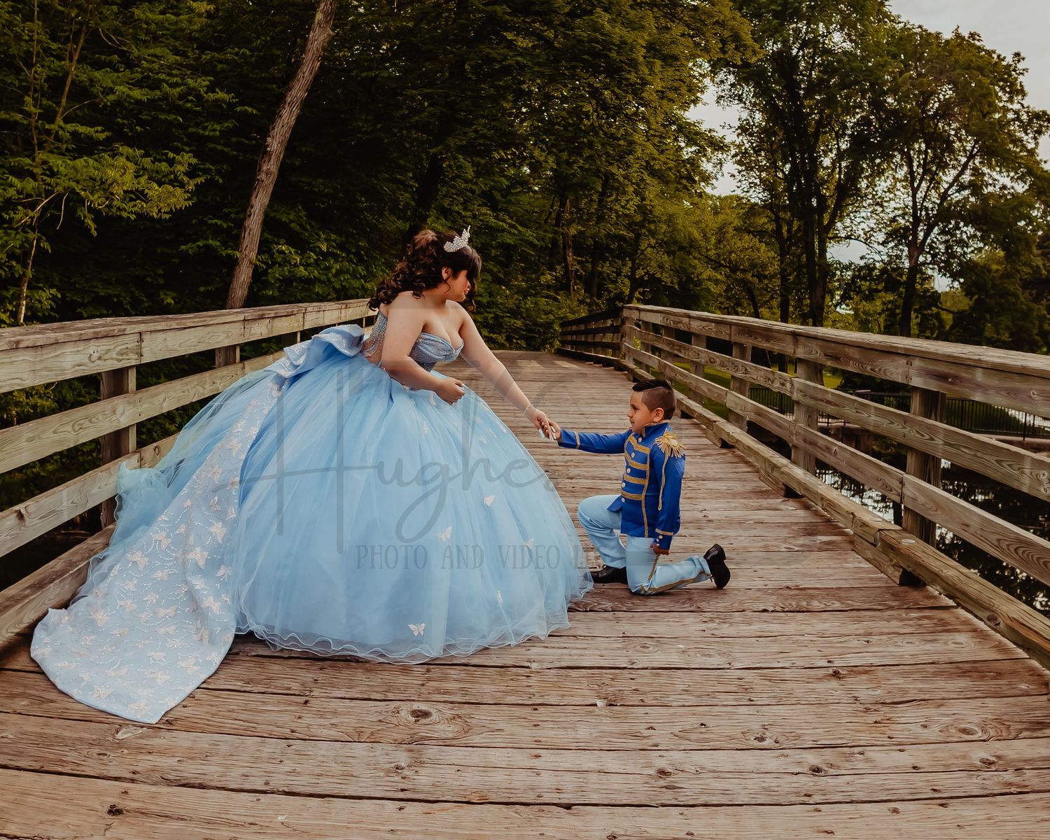 A light blue quinceañera dress worn on a wooden bridge with a small companion in blue kneeling beside.