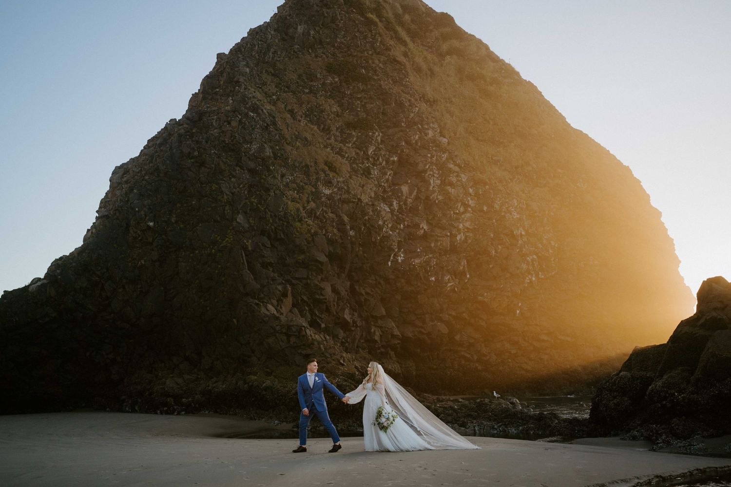 Silhouetted wedding couple holding hands against a golden sunset behind a large beach rock formation.