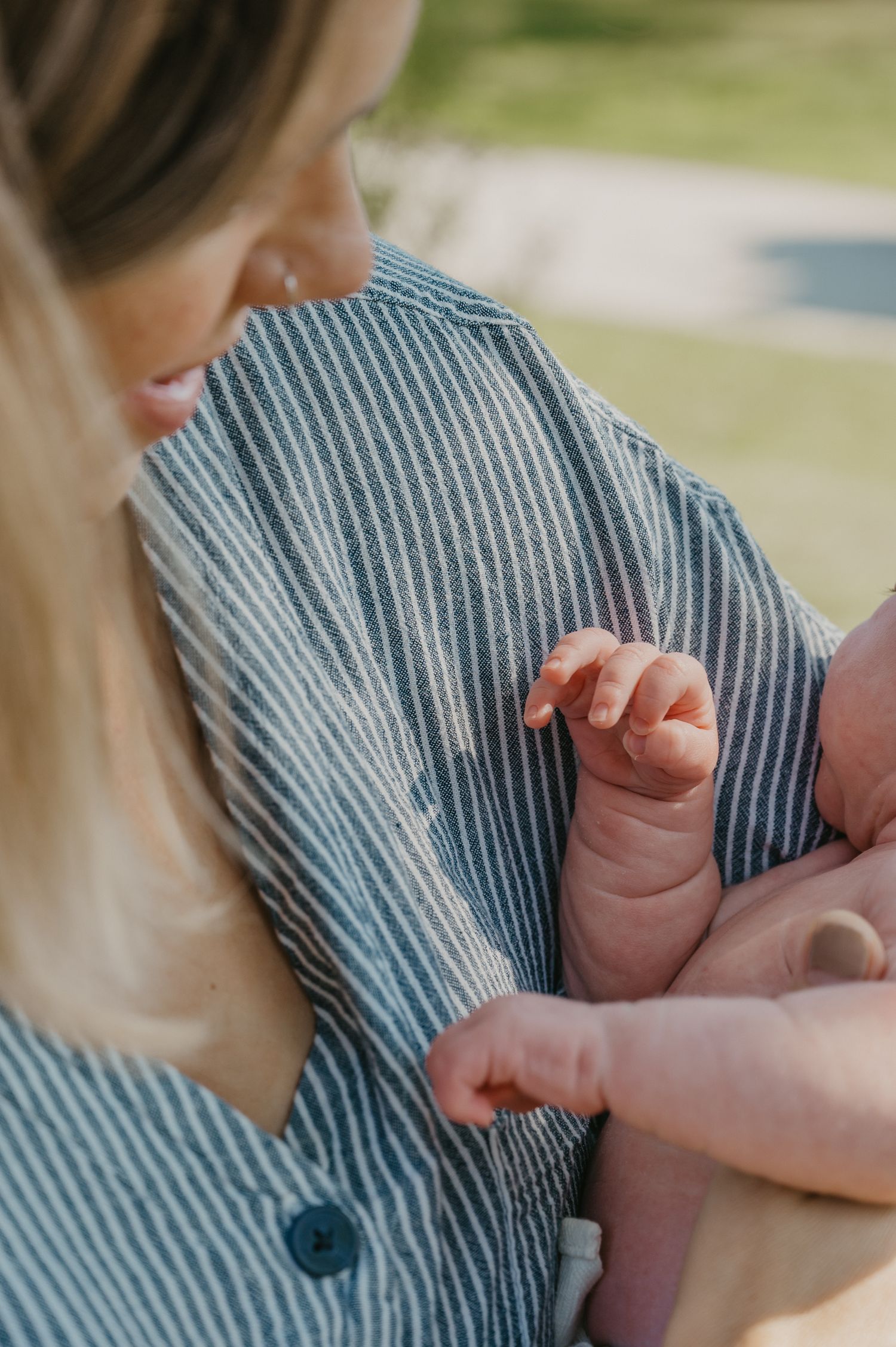 A mother in a striped dress tenderly holds her newborn baby's tiny hand outdoors.