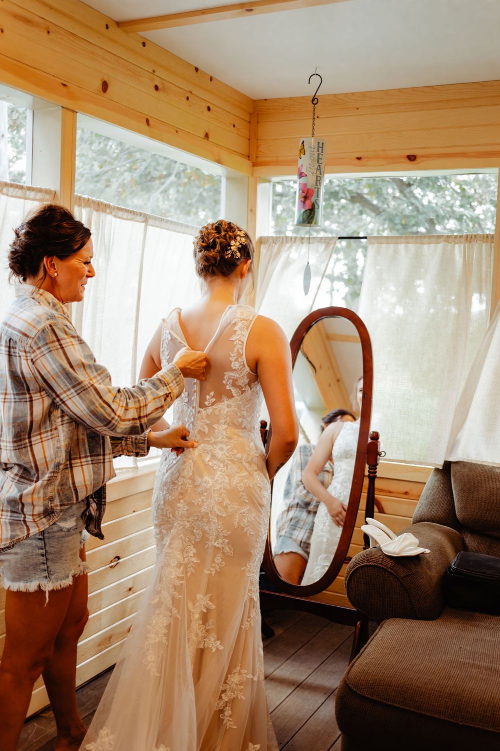 A person helps adjust a lace wedding dress in a sunlit wooden room with mirrors.