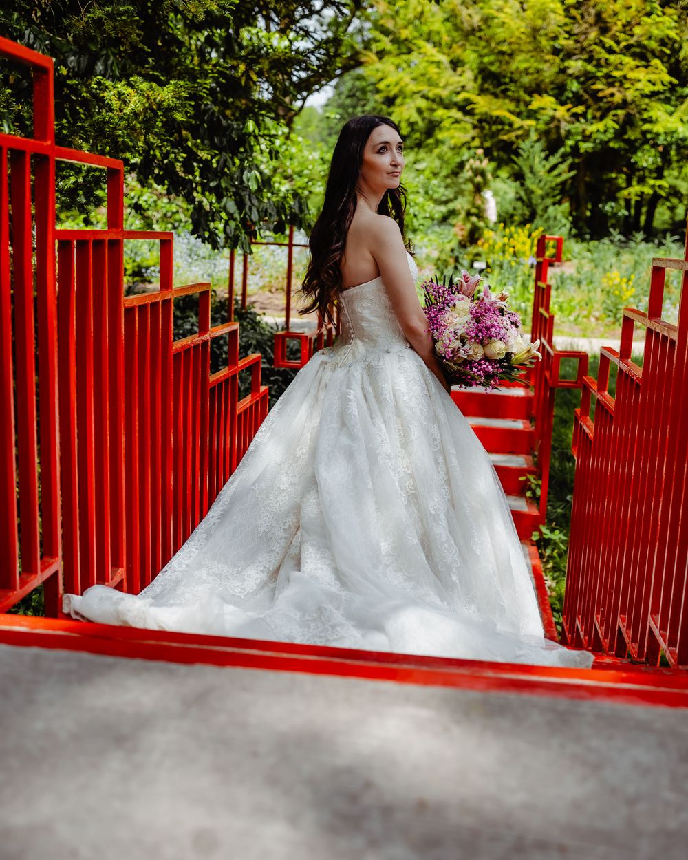 Bride in elegant ball gown poses on red bridge walkway with floral bouquet.