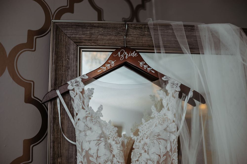 A delicate white lace wedding dress hangs on a wooden hanger against an ornate wall.