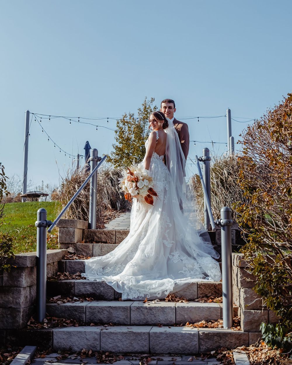 Wedding couple on stone steps with string lights and autumn foliage in background.