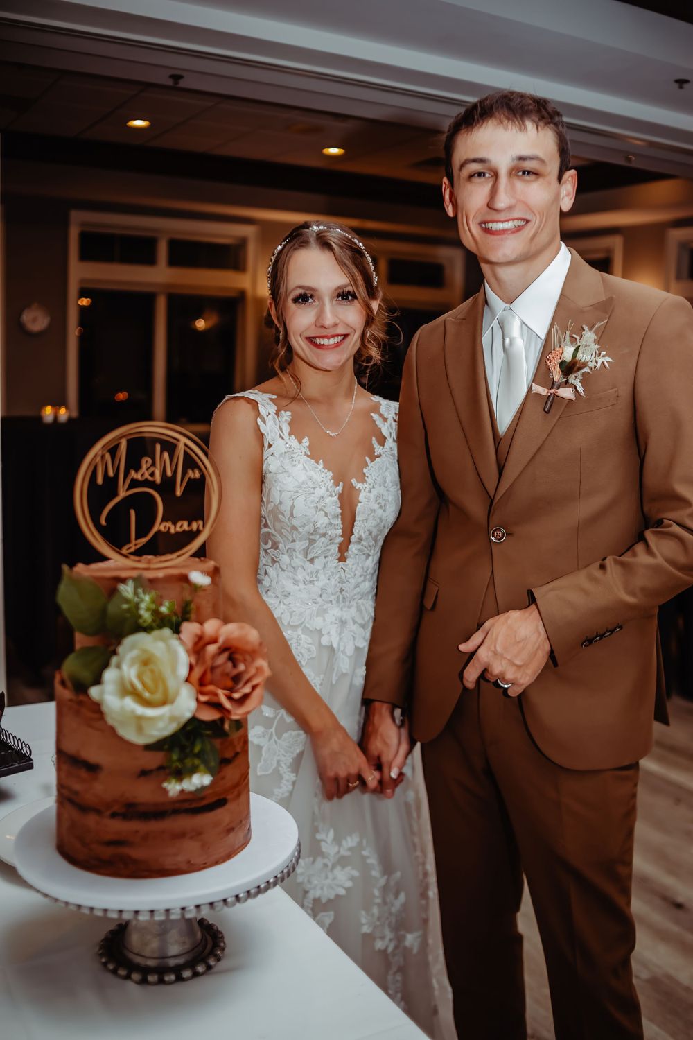 Wedding couple stands beside their rustic brown naked cake decorated with fresh flowers.
