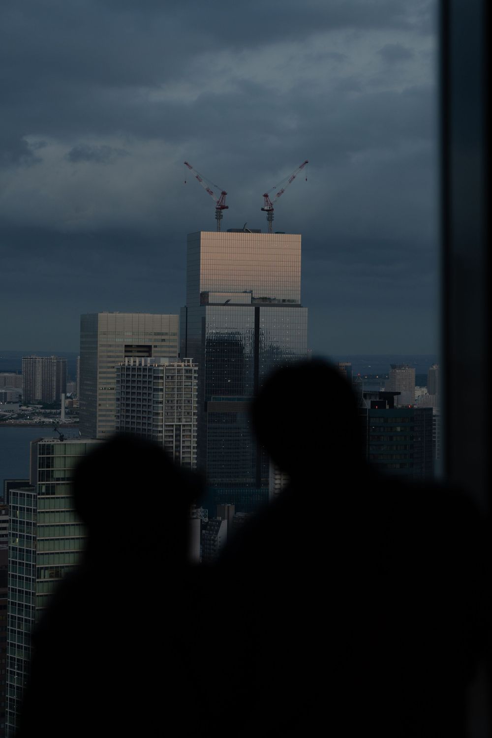 Silhouetted figures viewing a construction site with cranes against a stormy city skyline.