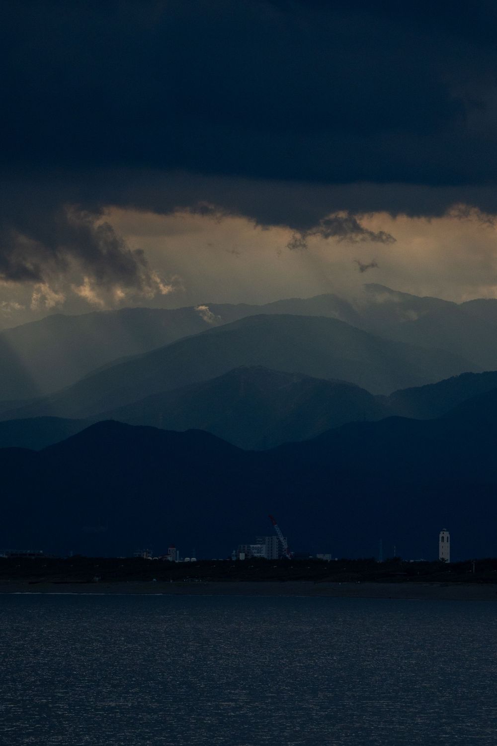 Dramatic mountain landscape at dusk with dark clouds and industrial buildings visible along the waterfront.