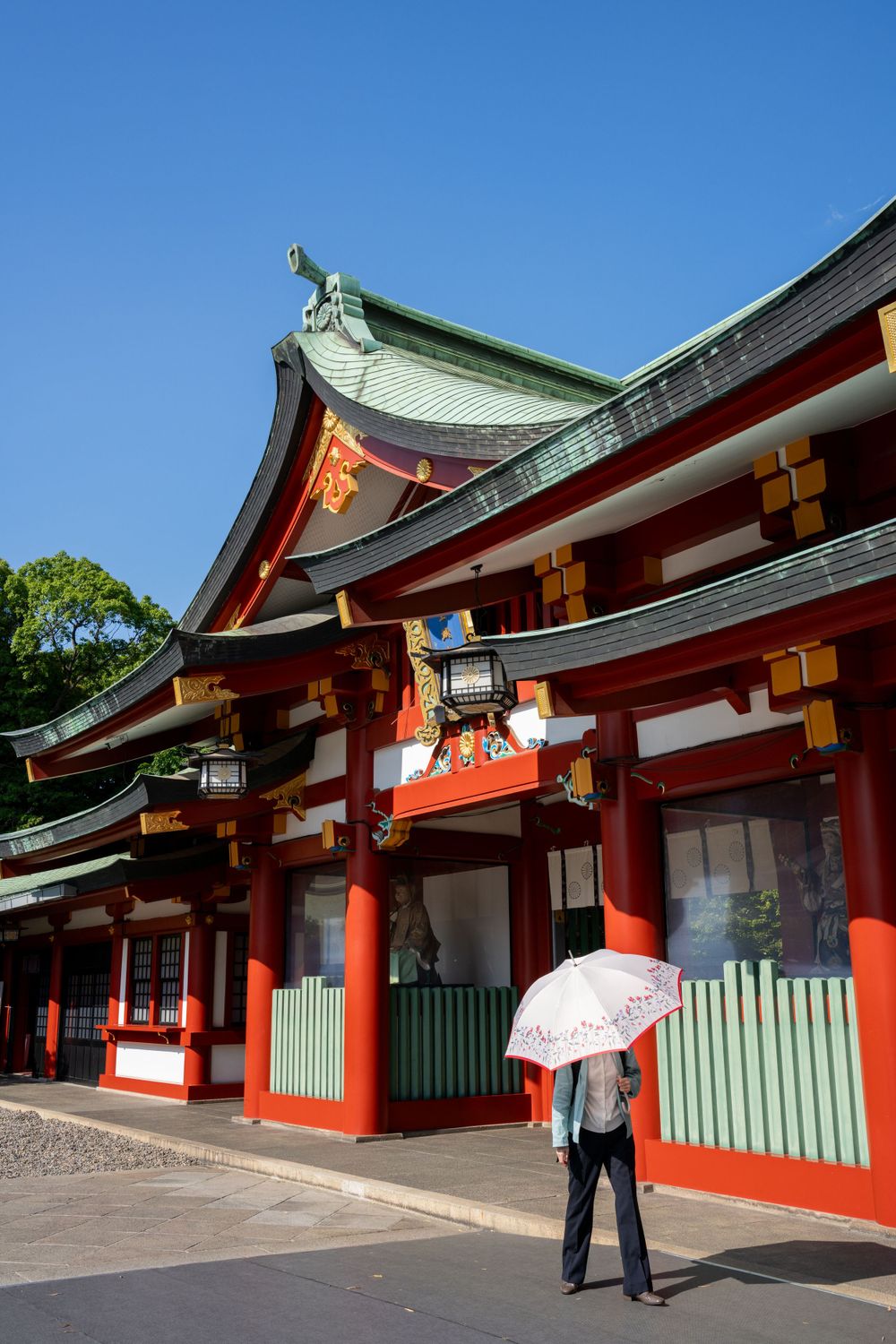 Traditional Japanese shrine with red pillars and ornate roof while someone holds a parasol on a sunny day.
