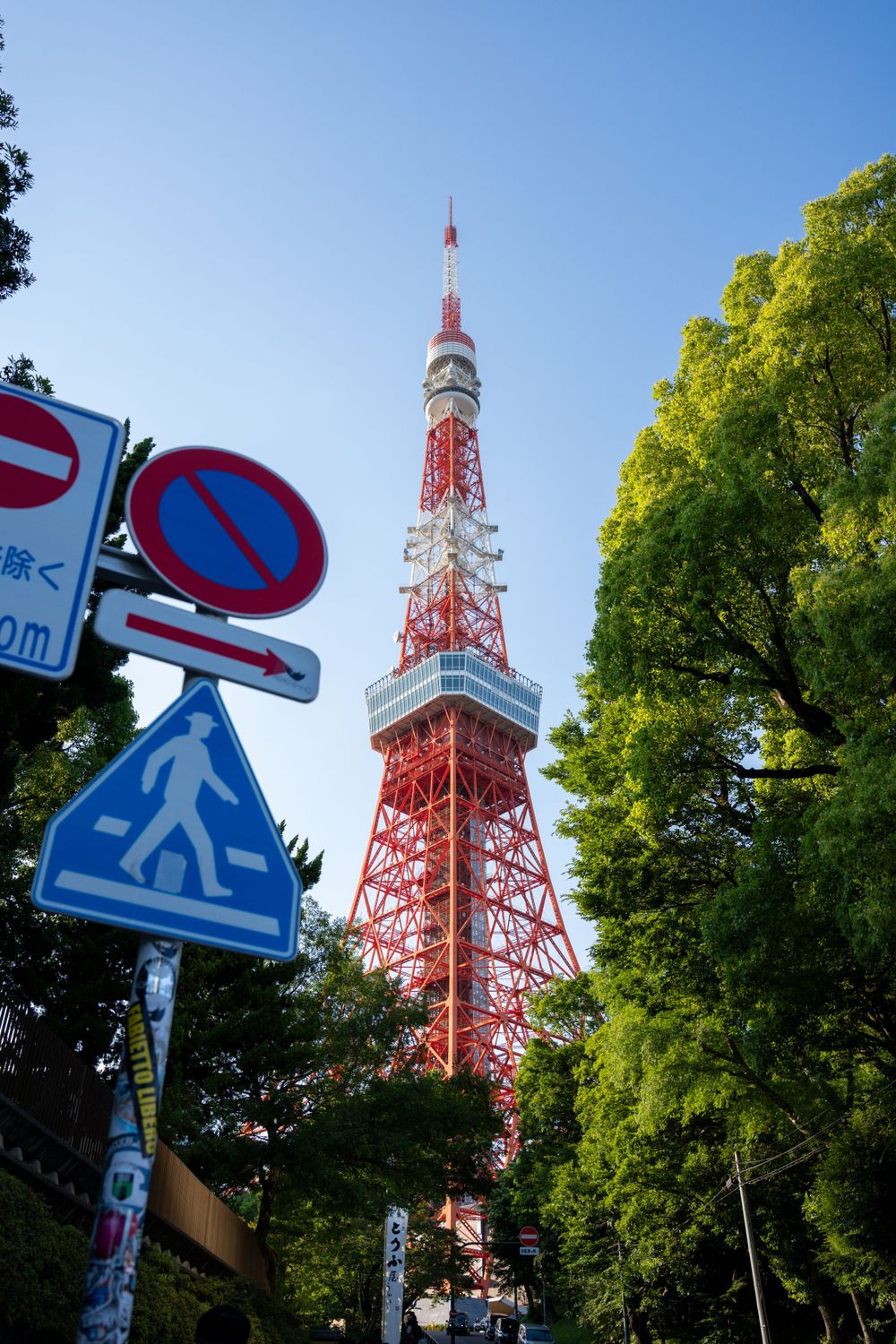 Tokyo Tower rises above traffic signs and green trees against a bright blue sky.