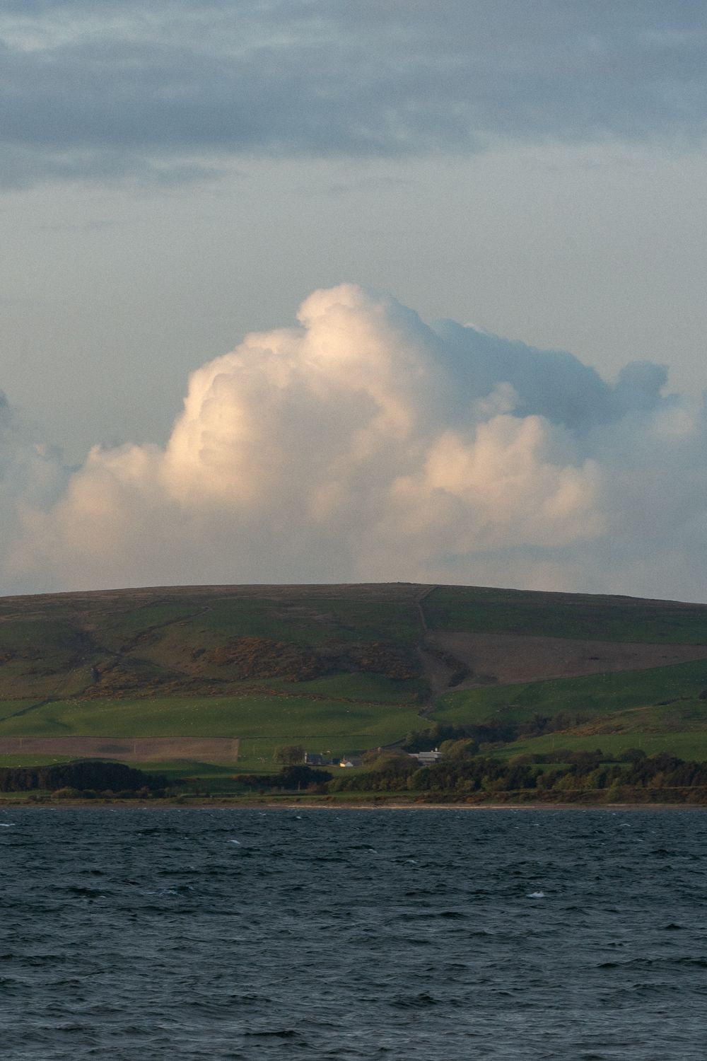 A dramatic cloudy sky looms over rolling green hills and dark waters along a rugged coastline.
