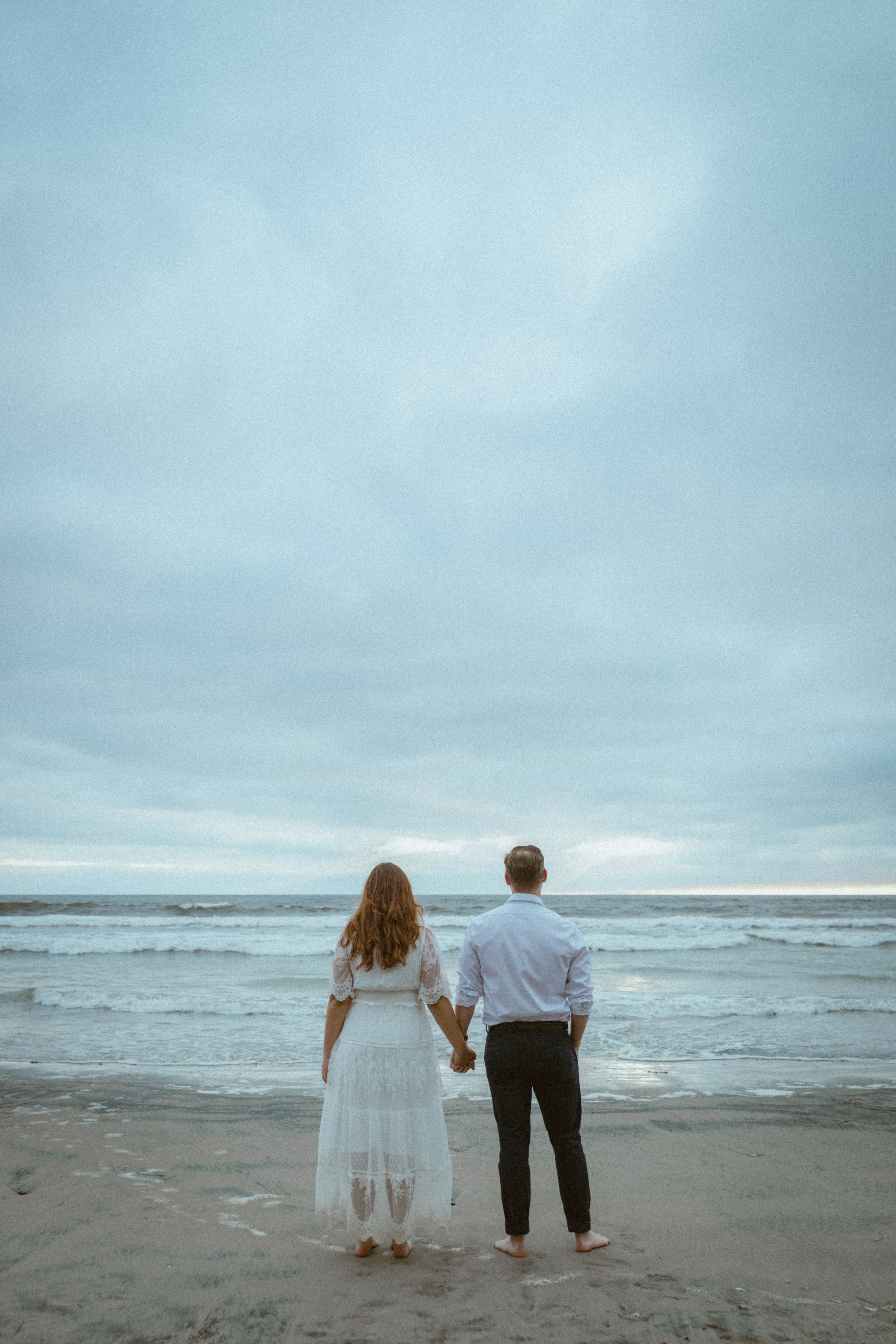 A couple stands hand in hand facing the ocean waves on an overcast beach.