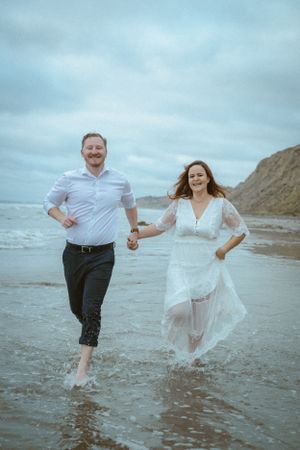 A couple runs joyfully along the beach with waves crashing behind them.