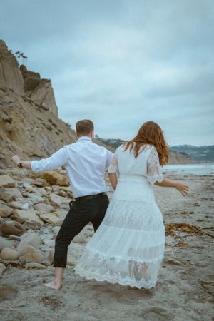 A playful couple dances on a rocky beach with cliffs in the background.