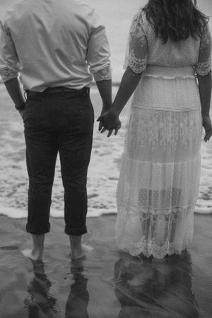 A couple stands barefoot in shallow ocean water wearing formal attire.