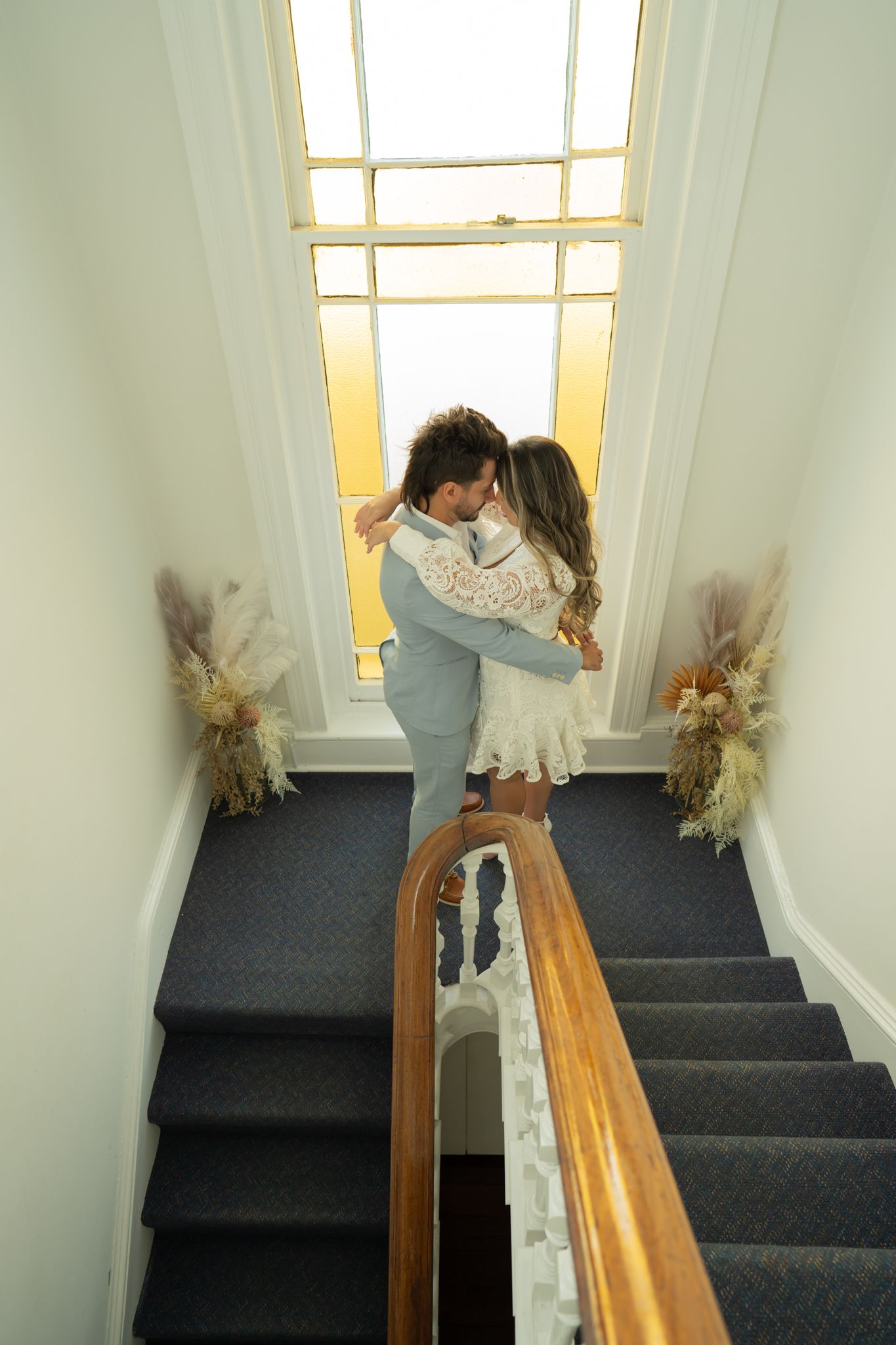Couple embraces near bright window on carpeted staircase landing with dried flower decor.