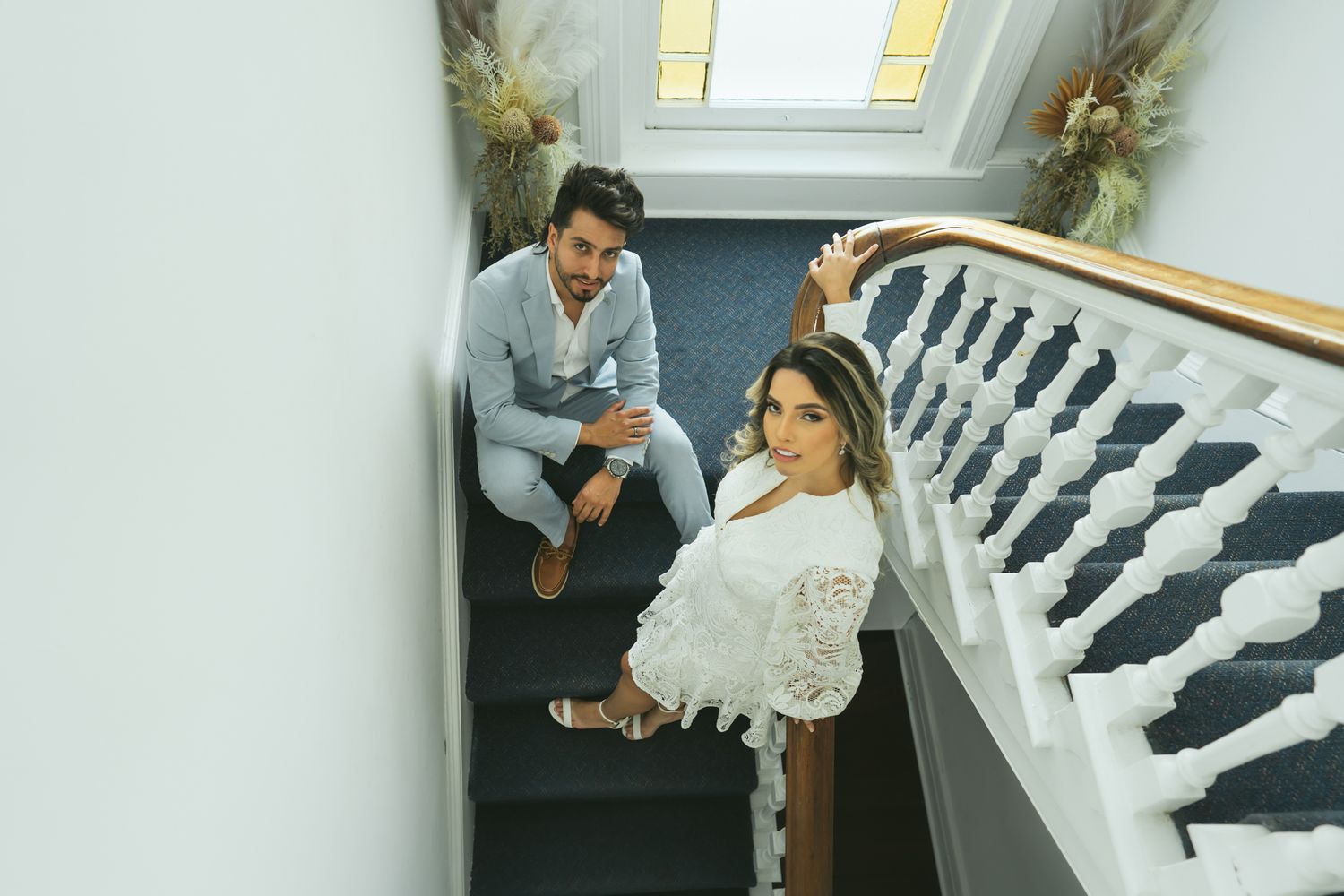 Couple sits together on carpeted stairs beside ornate white banister.