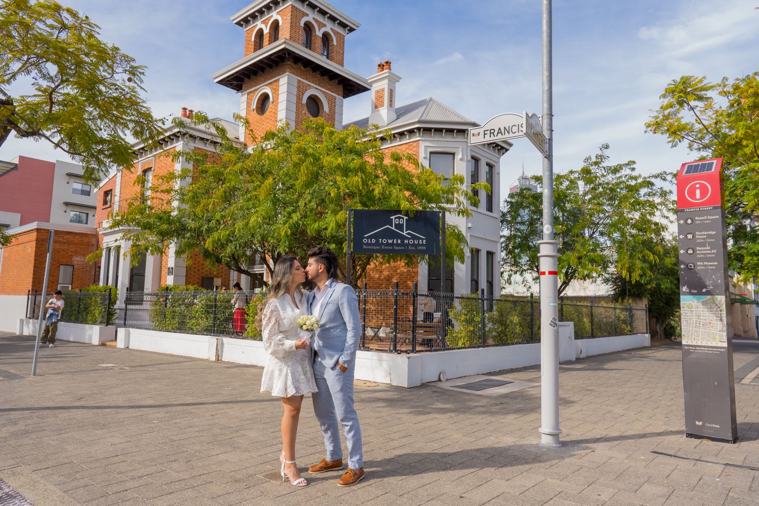 Couple poses outside historic brick building with clock tower on sunny day.