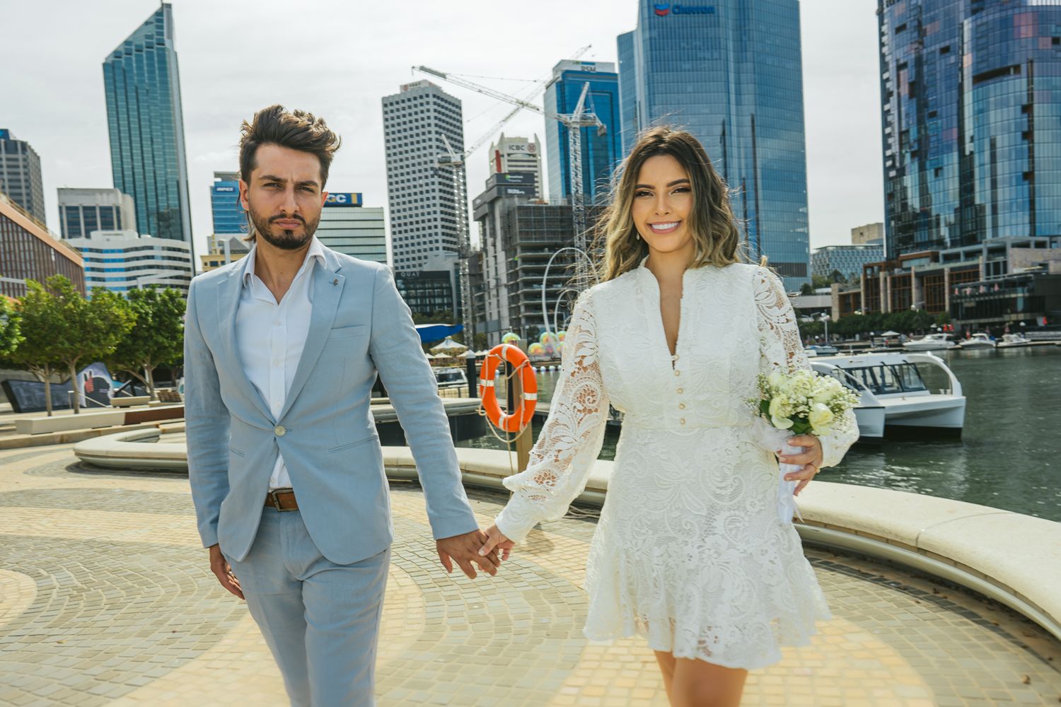 Stylish couple in light suits hold hands against modern city skyline backdrop.