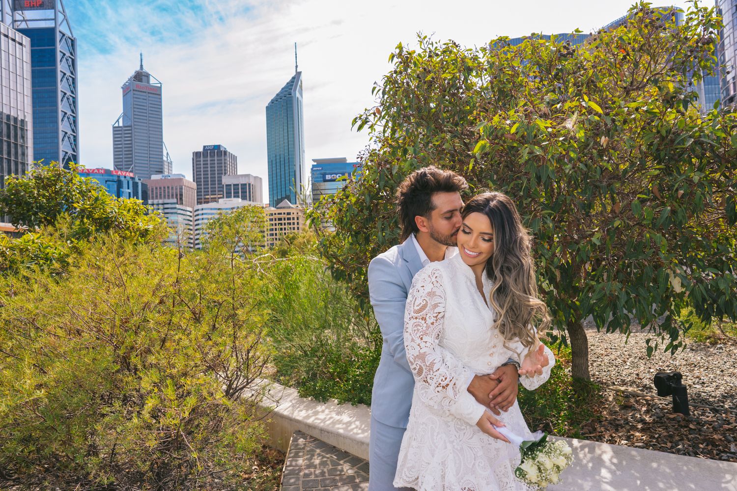 A couple embraces in white attire against Perth's modern cityscape with skyscrapers and autumn foliage.