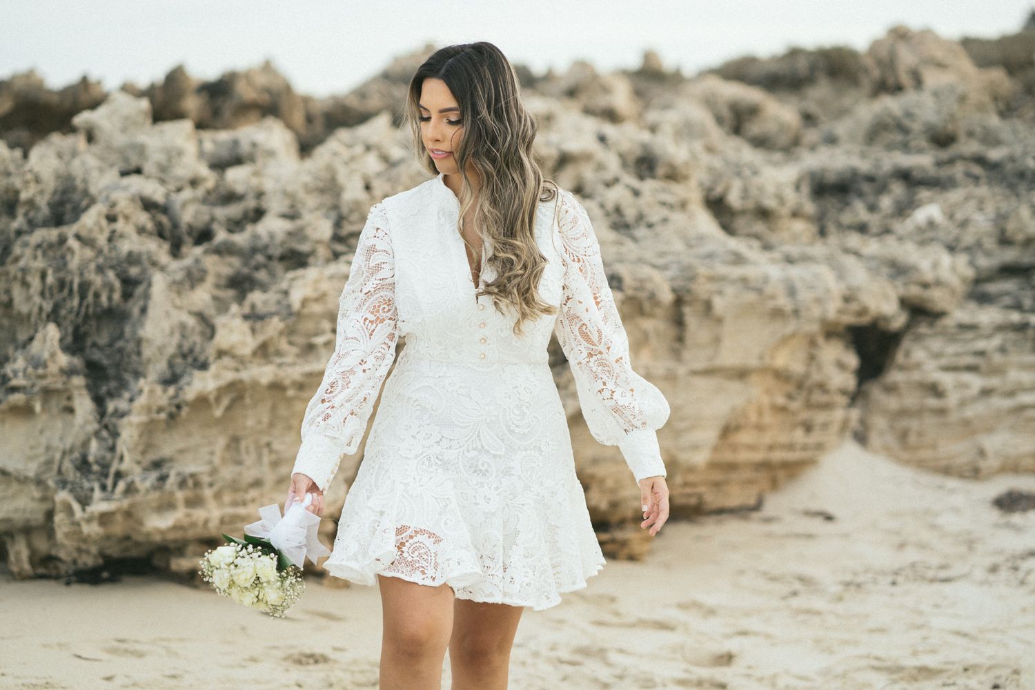 A person in white lace dress holds bouquet against coastal rock formations.