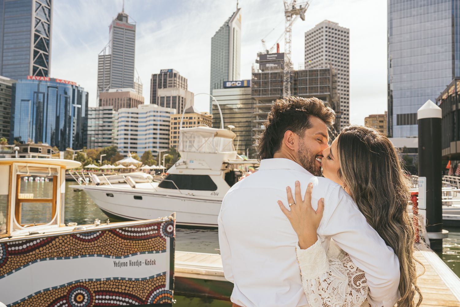 An intimate embrace on a deck with Perth's urban skyline and boats in the background.