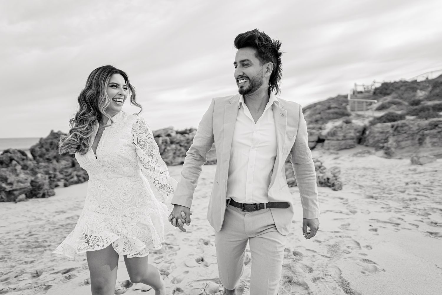 Black and white portrait of couple walking hand in hand on rocky beach.