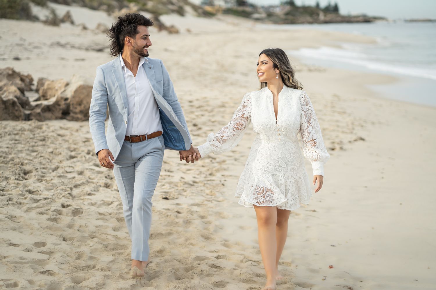 Couple strolling together along scenic beach wearing coordinated light colored outfits.