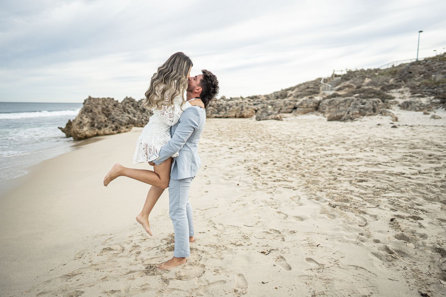 A joyful couple in denim sharing a playful lift embrace on a rocky beach.