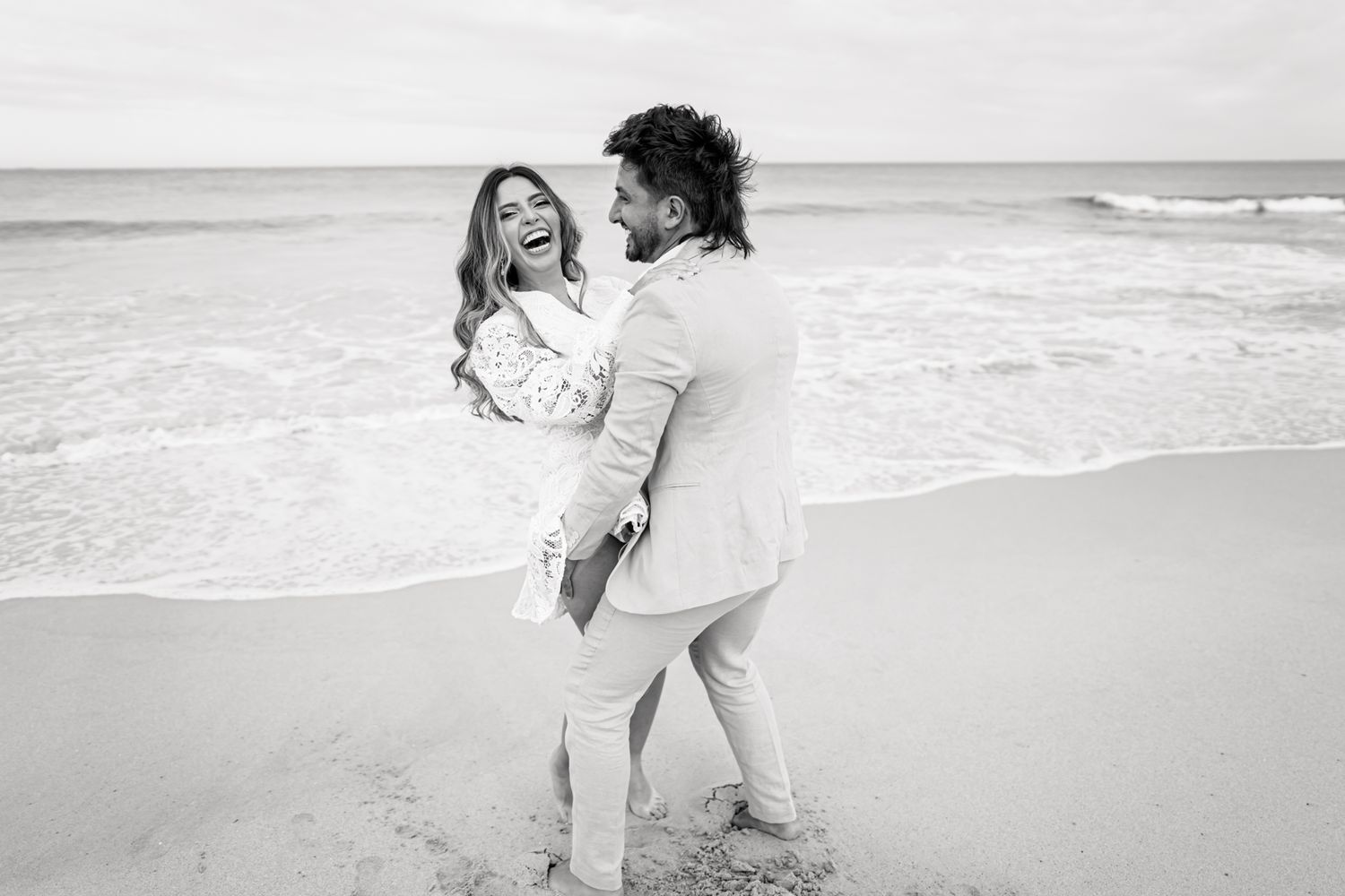 A romantic couple laughing and embracing on a beach in black and white photography.