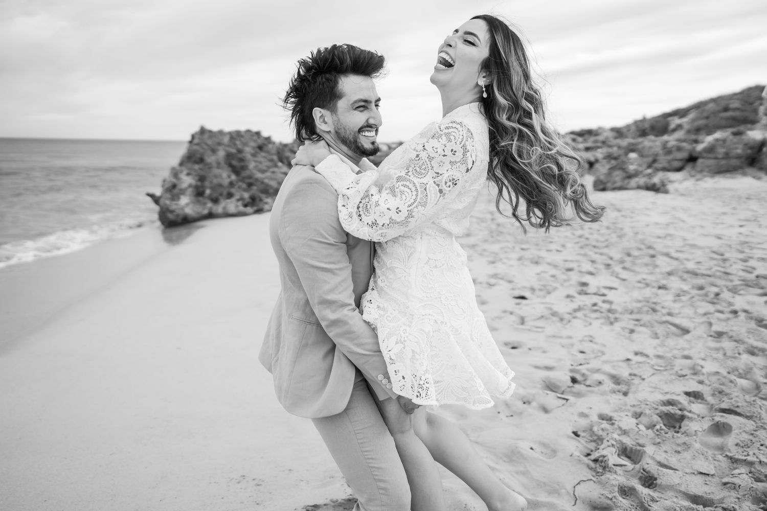 A happy couple dancing and laughing together on a scenic beach with rocky cliffs.