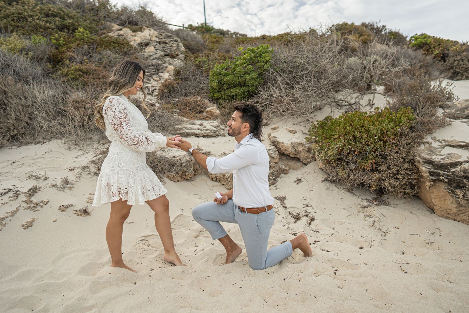 A romantic beach proposal with coastal vegetation and dramatic clouds in the background.
