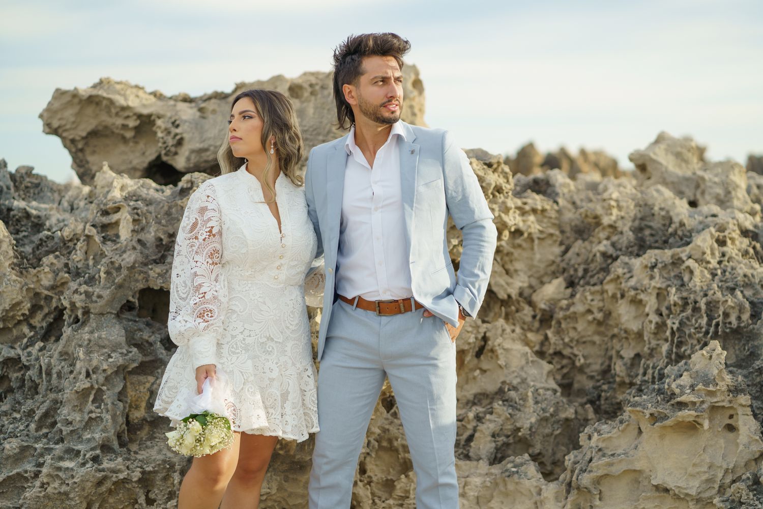 Stylish couple in light attire pose against dramatic rocky coastal backdrop.