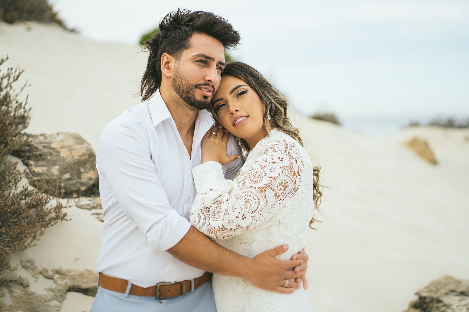 A loving couple in white attire embracing on sandy dunes with coastal vegetation.